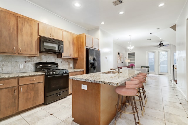 kitchen with ceiling fan with notable chandelier, light stone counters, sink, black appliances, and a center island with sink