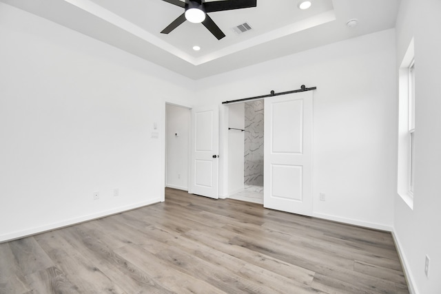 unfurnished bedroom with ceiling fan, a raised ceiling, a barn door, ensuite bathroom, and light wood-type flooring