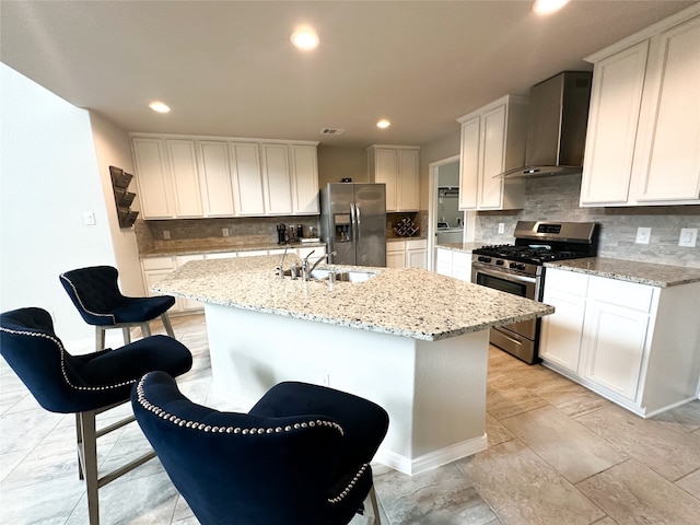 kitchen with wall chimney range hood, stainless steel appliances, an island with sink, and white cabinetry