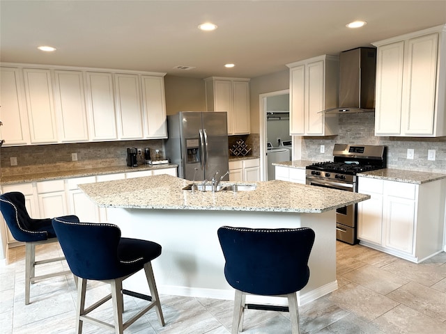 kitchen featuring appliances with stainless steel finishes, white cabinetry, an island with sink, and wall chimney range hood