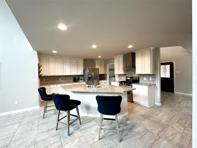 kitchen with wall chimney range hood, white cabinets, appliances with stainless steel finishes, and a kitchen island with sink