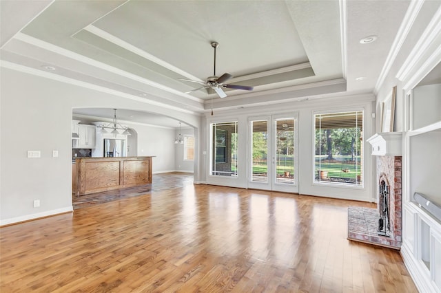 unfurnished living room featuring a raised ceiling, ornamental molding, ceiling fan with notable chandelier, and a brick fireplace