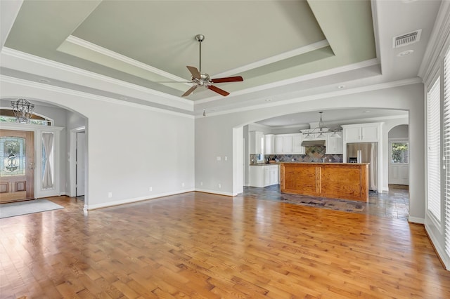 unfurnished living room with ceiling fan with notable chandelier, light hardwood / wood-style floors, crown molding, and a tray ceiling