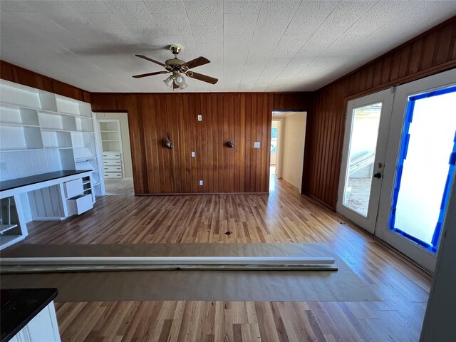 unfurnished living room with french doors, ceiling fan, wooden walls, and light hardwood / wood-style floors