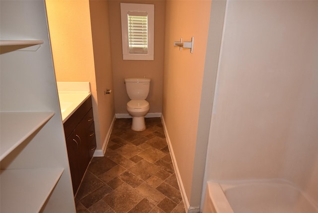 bathroom with baseboards, toilet, a tub to relax in, stone tile flooring, and vanity