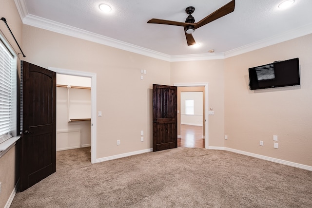 unfurnished bedroom with a spacious closet, a closet, crown molding, carpet flooring, and ceiling fan