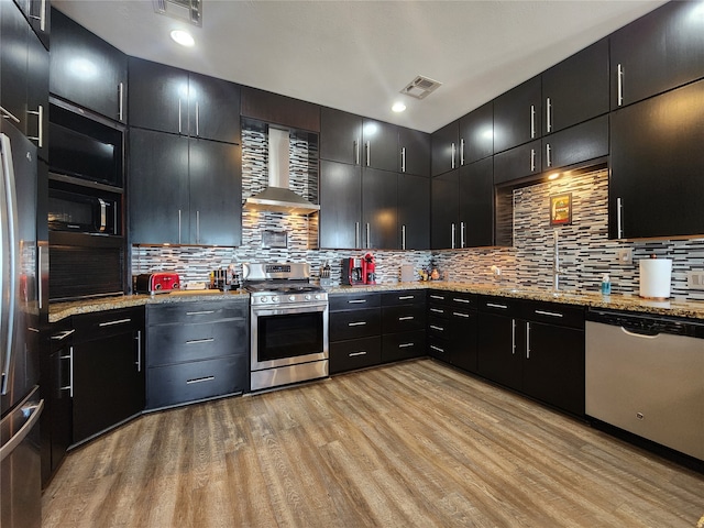 kitchen featuring light stone countertops, appliances with stainless steel finishes, tasteful backsplash, wall chimney exhaust hood, and light hardwood / wood-style flooring