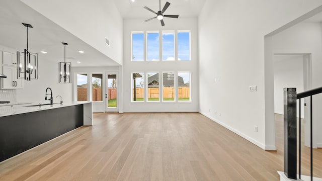 unfurnished living room with light hardwood / wood-style flooring, sink, ceiling fan with notable chandelier, and a high ceiling