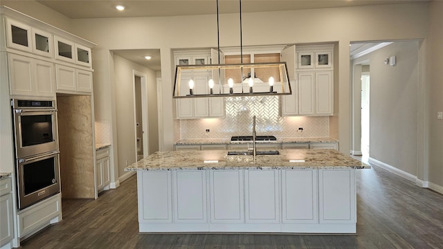 kitchen featuring stainless steel double oven, decorative light fixtures, a kitchen island with sink, light stone countertops, and sink