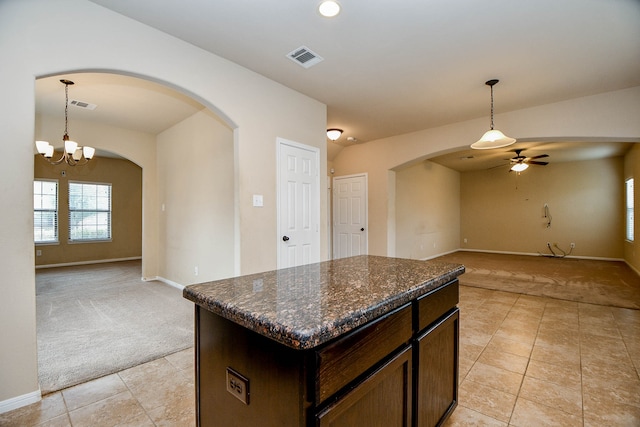 kitchen with a kitchen island, ceiling fan with notable chandelier, light carpet, dark brown cabinetry, and dark stone counters