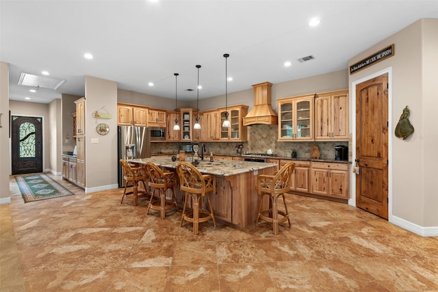 kitchen featuring backsplash, premium range hood, an island with sink, appliances with stainless steel finishes, and light stone counters