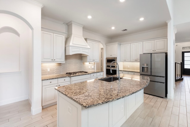 kitchen with custom exhaust hood, stainless steel appliances, white cabinetry, and a kitchen island with sink