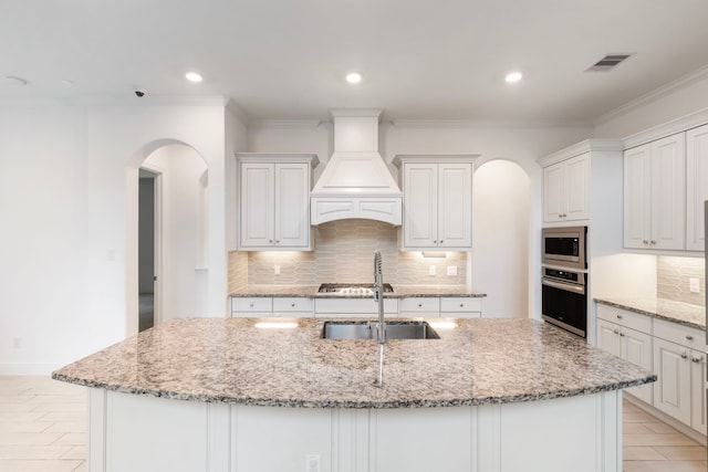 kitchen featuring an island with sink, stainless steel appliances, light stone counters, and premium range hood