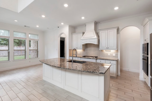 kitchen featuring a center island with sink, premium range hood, sink, dark stone countertops, and white cabinets