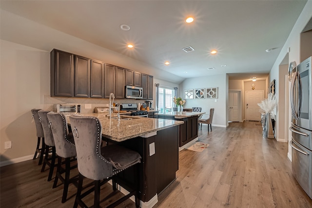 kitchen featuring light stone countertops, appliances with stainless steel finishes, a kitchen bar, dark brown cabinetry, and light wood-type flooring