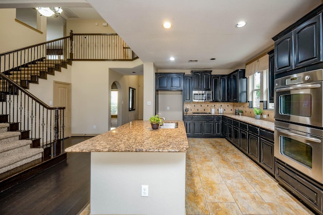 kitchen with stainless steel appliances, backsplash, a kitchen island with sink, a sink, and light stone countertops