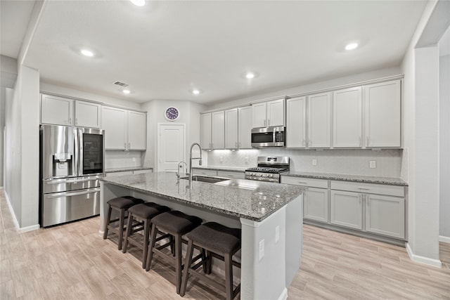 kitchen with a kitchen island with sink, sink, light stone countertops, appliances with stainless steel finishes, and light hardwood / wood-style floors