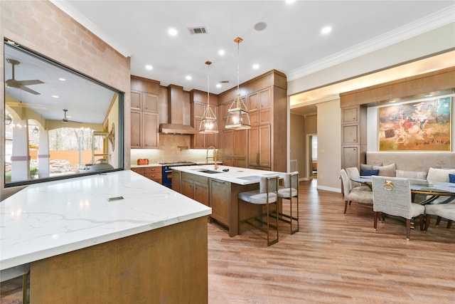 kitchen with light wood-type flooring, ceiling fan, a breakfast bar, wall chimney exhaust hood, and a center island with sink