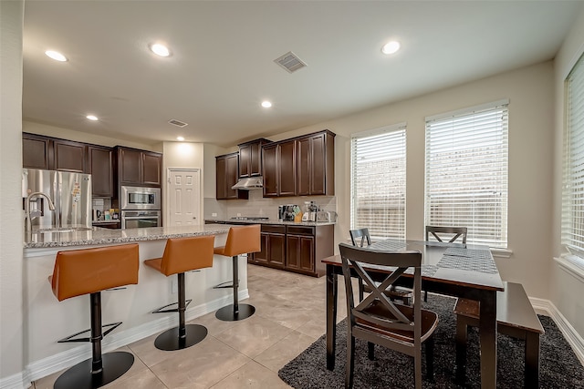 kitchen with dark brown cabinets, light tile patterned floors, stainless steel appliances, decorative backsplash, and light stone counters