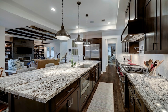 kitchen featuring a large fireplace, light stone counters, sink, built in appliances, and a center island with sink