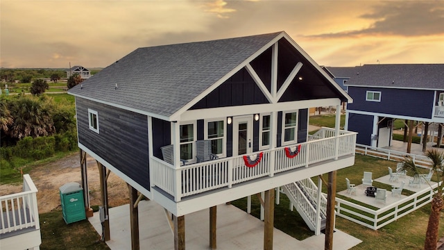 back of house at dusk with roof with shingles, a porch, stairway, board and batten siding, and fence