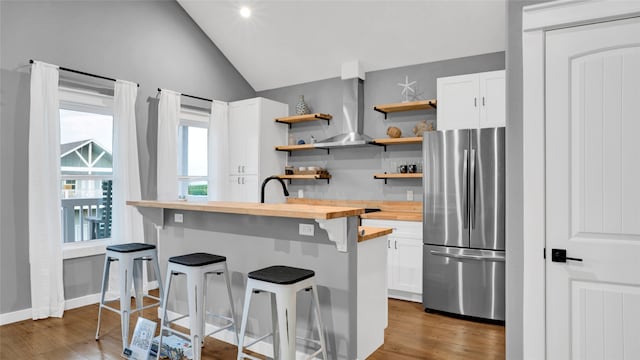 kitchen with vaulted ceiling, a kitchen breakfast bar, stainless steel fridge, butcher block counters, and wall chimney range hood