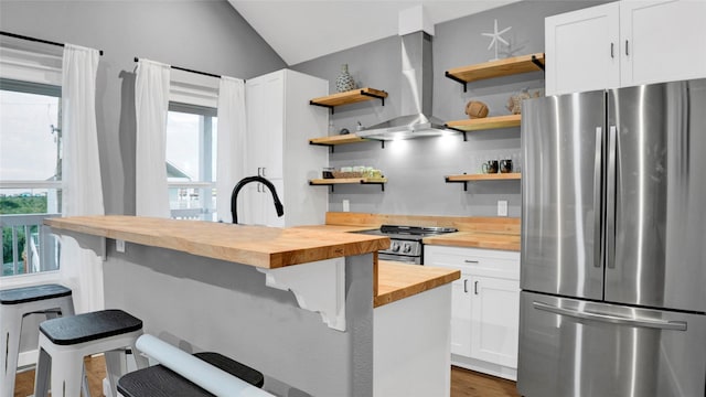 kitchen featuring a breakfast bar area, butcher block countertops, wall chimney range hood, appliances with stainless steel finishes, and open shelves