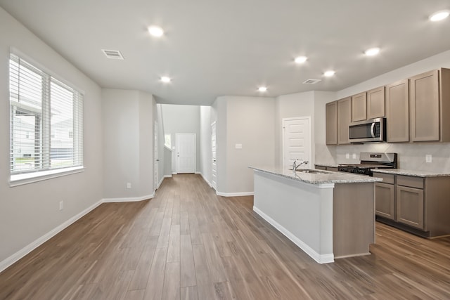 kitchen featuring wood-type flooring, appliances with stainless steel finishes, a kitchen island with sink, sink, and light stone counters