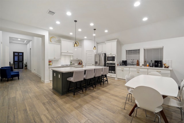 kitchen featuring light hardwood / wood-style flooring, stainless steel appliances, decorative light fixtures, a center island with sink, and white cabinets