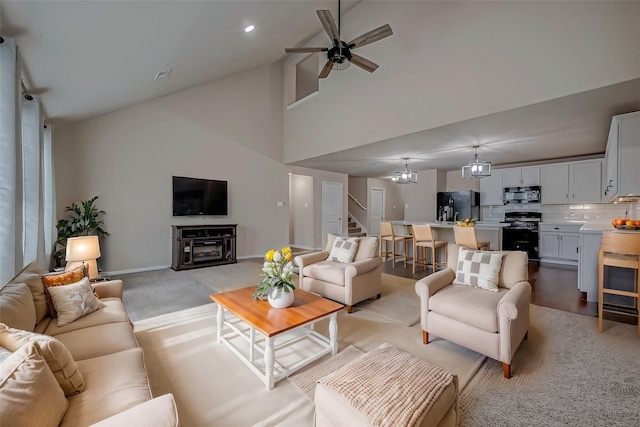 living area with ceiling fan with notable chandelier, light colored carpet, a high ceiling, baseboards, and stairs