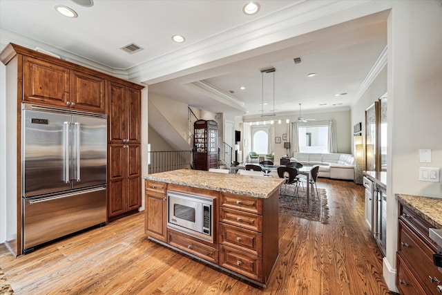 kitchen with light wood-type flooring, ornamental molding, built in appliances, a center island, and light stone countertops