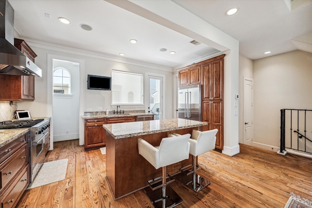 kitchen with light wood-type flooring, high end appliances, a kitchen island, sink, and wall chimney exhaust hood