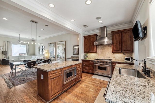 kitchen featuring wall chimney exhaust hood, light hardwood / wood-style floors, an island with sink, stainless steel appliances, and sink