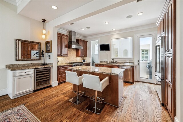 kitchen featuring a kitchen island, wine cooler, appliances with stainless steel finishes, wall chimney range hood, and light wood-type flooring