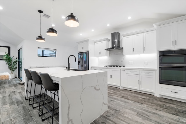 kitchen with appliances with stainless steel finishes, light hardwood / wood-style floors, a kitchen island with sink, white cabinetry, and wall chimney range hood