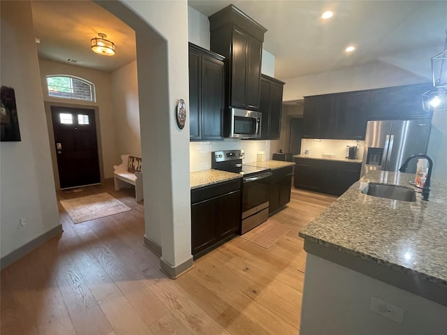 kitchen with a sink, dark cabinetry, light wood-style floors, appliances with stainless steel finishes, and light stone countertops