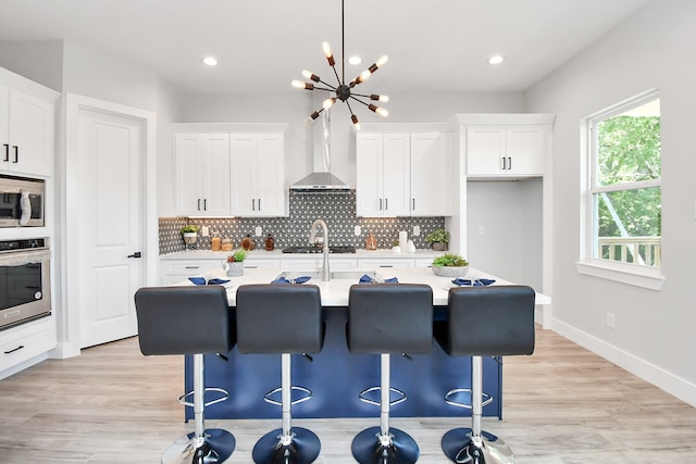 kitchen featuring a center island with sink, white cabinetry, wall chimney range hood, and stainless steel appliances