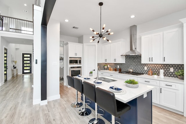 kitchen featuring wall chimney exhaust hood, white cabinetry, stainless steel appliances, sink, and a kitchen island with sink