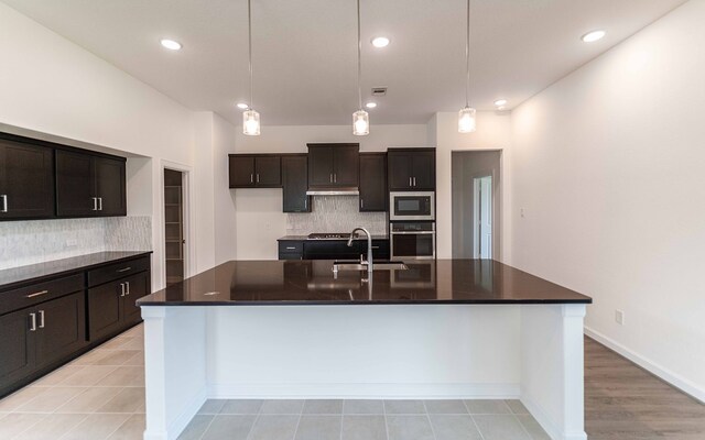 kitchen with an island with sink, hanging light fixtures, dark brown cabinets, appliances with stainless steel finishes, and tasteful backsplash