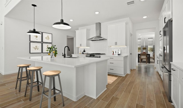 kitchen with wall chimney exhaust hood, a kitchen island with sink, sink, light hardwood / wood-style flooring, and white cabinets