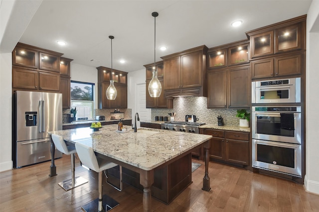 kitchen with a breakfast bar, light stone counters, stainless steel appliances, a center island with sink, and dark hardwood / wood-style flooring