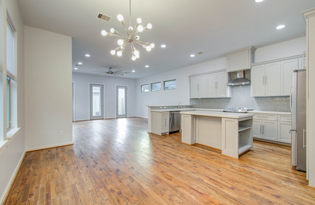 kitchen with ceiling fan with notable chandelier, stainless steel appliances, a center island, light hardwood / wood-style flooring, and wall chimney range hood