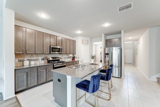 kitchen with appliances with stainless steel finishes, light stone counters, sink, an island with sink, and a breakfast bar