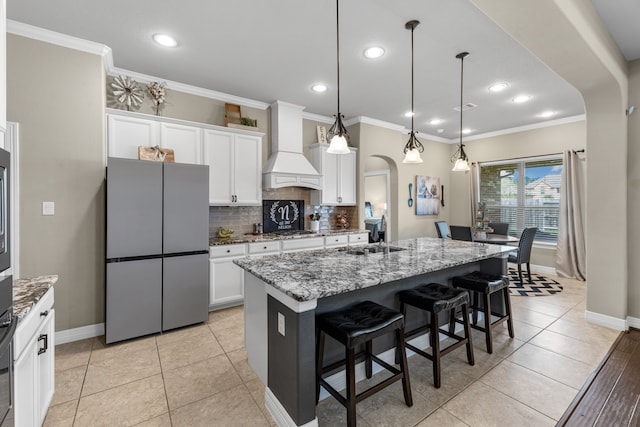 kitchen with a center island with sink, dark stone countertops, stainless steel appliances, decorative light fixtures, and premium range hood