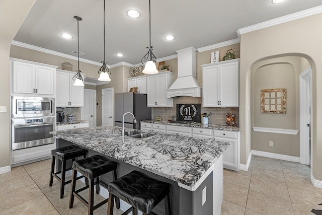 kitchen with custom exhaust hood, decorative light fixtures, a kitchen island with sink, white cabinetry, and appliances with stainless steel finishes