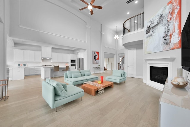 living area featuring ceiling fan with notable chandelier, a towering ceiling, stairs, light wood-type flooring, and a glass covered fireplace