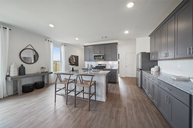 kitchen with gray cabinetry, dark hardwood / wood-style flooring, appliances with stainless steel finishes, an island with sink, and light stone counters