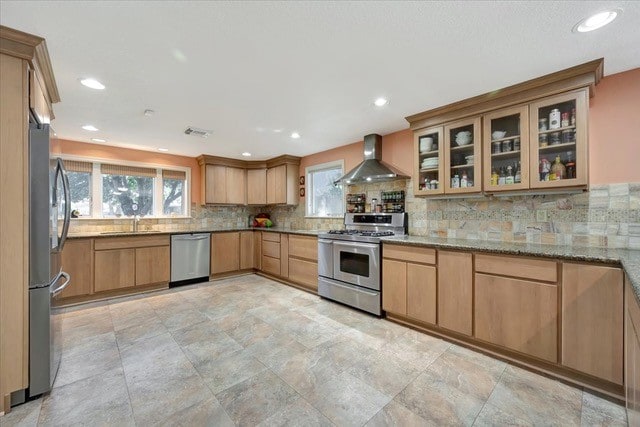 kitchen featuring wall chimney range hood, light stone counters, decorative backsplash, light brown cabinetry, and appliances with stainless steel finishes