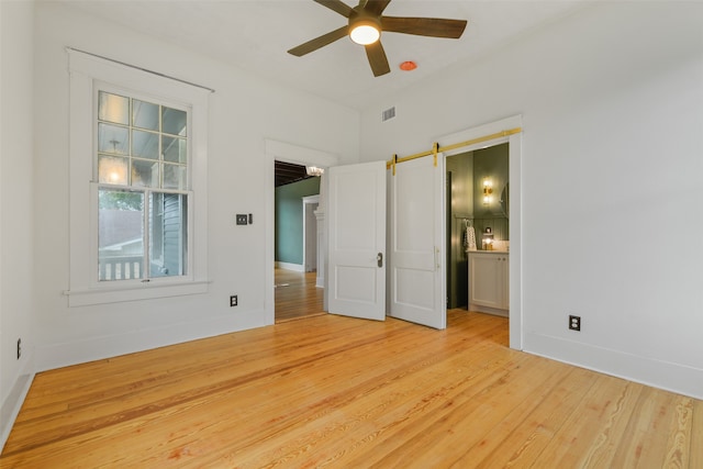 unfurnished bedroom with ensuite bath, light hardwood / wood-style floors, ceiling fan, and a barn door