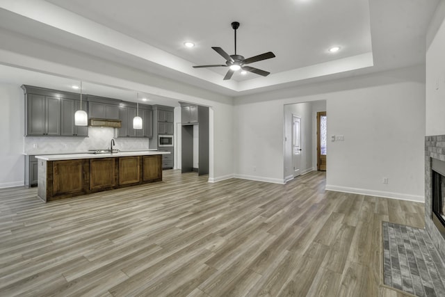 unfurnished living room with a tile fireplace, ceiling fan, sink, light hardwood / wood-style floors, and a tray ceiling